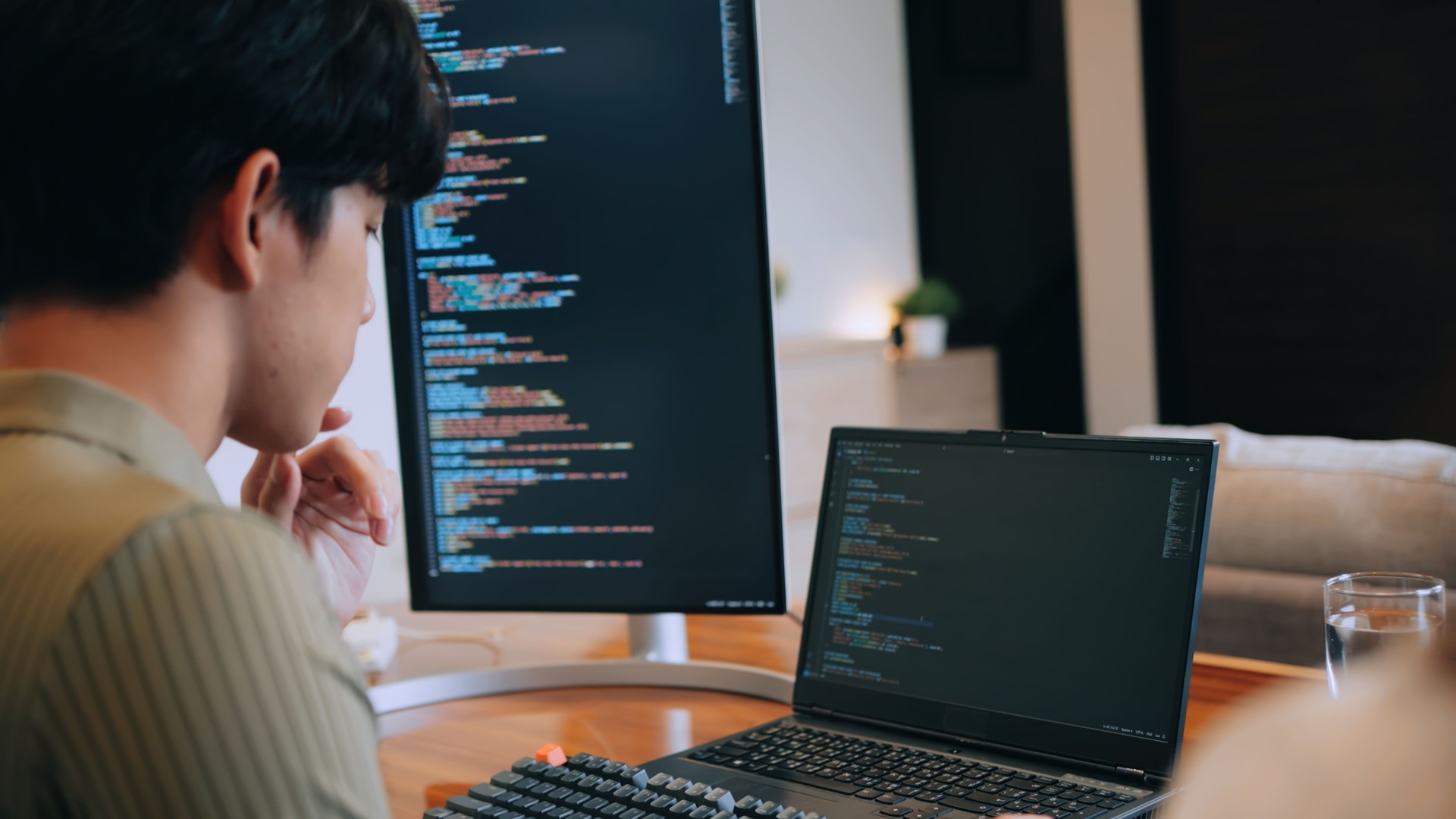 A developer coding on a laptop with a dual-screen setup in a home office. A relatable scene of focus, productivity, and modern technology. Over the shoulder view